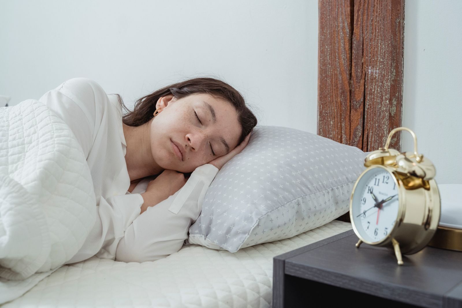 Young calm female with dark hair lying on side with hand under head on white comfortable bed under blanket while sleeping in morning time
