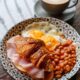 A hearty plate of English breakfast with eggs, beans, ham croissant, and coffee on a patterned plate.
