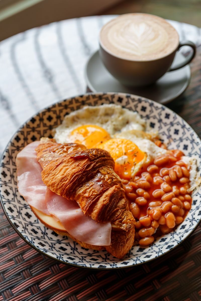 A hearty plate of English breakfast with eggs, beans, ham croissant, and coffee on a patterned plate.