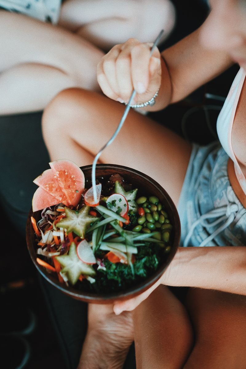 From above of crop anonymous young woman in summer clothes sitting with crossed legs and enjoying healthy salad bowl