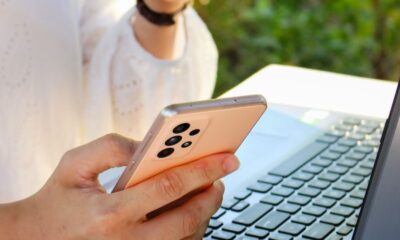 Person using a smartphone while drinking coffee near a laptop outdoors, showcasing multitasking with technology.