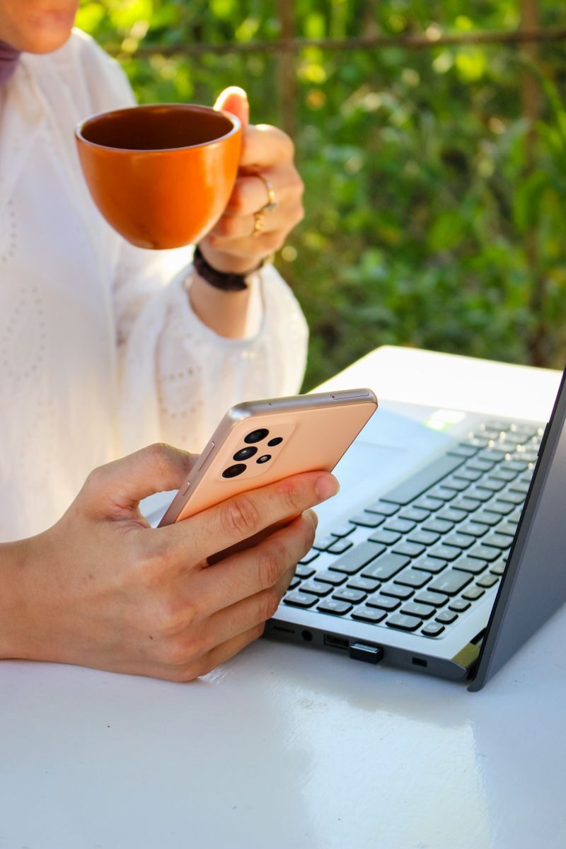 Person using a smartphone while drinking coffee near a laptop outdoors, showcasing multitasking with technology.
