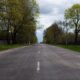 A long, empty road bordered by lush trees in a peaceful spring countryside setting.