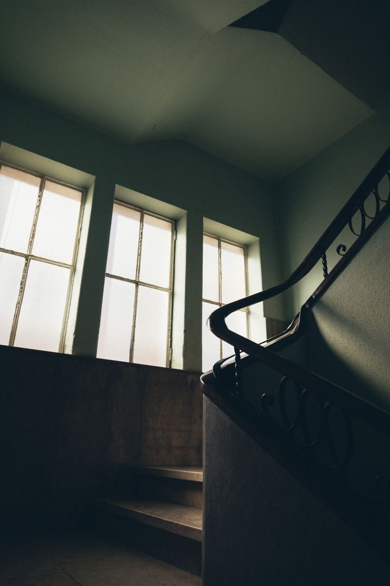 Dimly lit vintage staircase with iron handrail and large windows, creating a nostalgic atmosphere.
