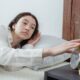 Sleepy young brunette female in white sleepwear resting in comfortable bed and waking up with alarm clock at home in morning time