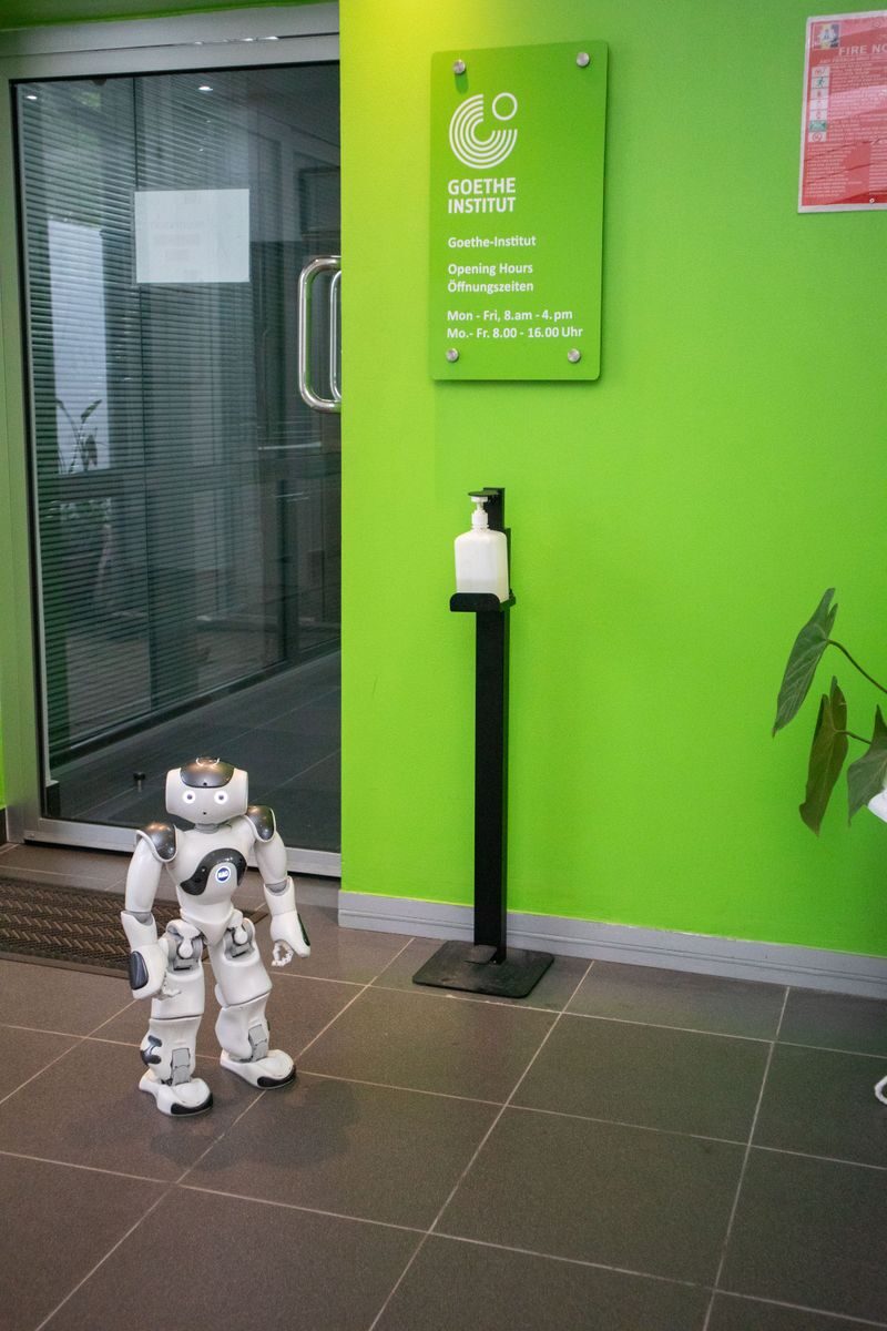 A robot stands near a sanitizer dispenser inside the Goethe Institut with a green wall backdrop.