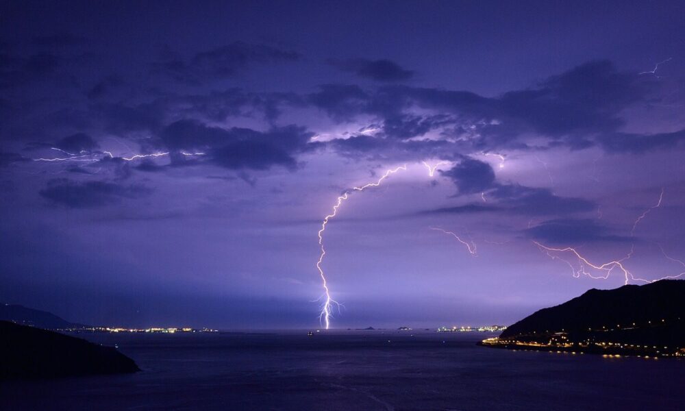 Spectacular lightning strikes illuminate a nighttime ocean view with dramatic clouds and city lights.