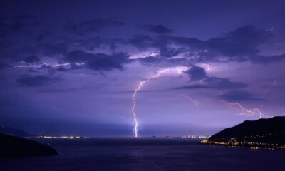 Spectacular lightning strikes illuminate a nighttime ocean view with dramatic clouds and city lights.