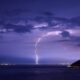 Spectacular lightning strikes illuminate a nighttime ocean view with dramatic clouds and city lights.