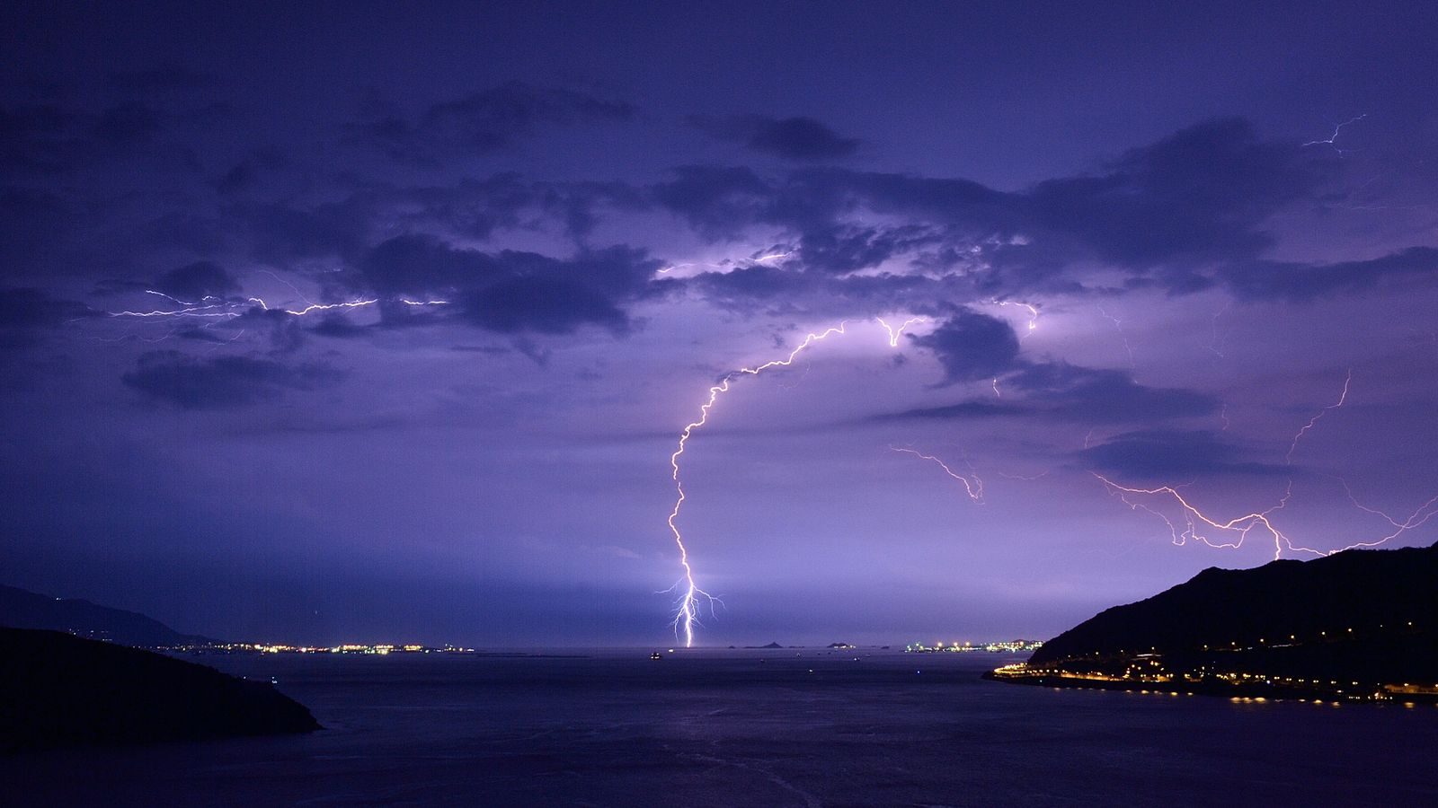 Spectacular lightning strikes illuminate a nighttime ocean view with dramatic clouds and city lights.