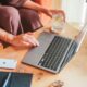 Woman using a laptop at home with a glass of lemon water, smartphone, and notebook on the table.