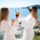 Two women in white robes toast with drinks on a sunny day by the sea, exuding relaxation and leisure.