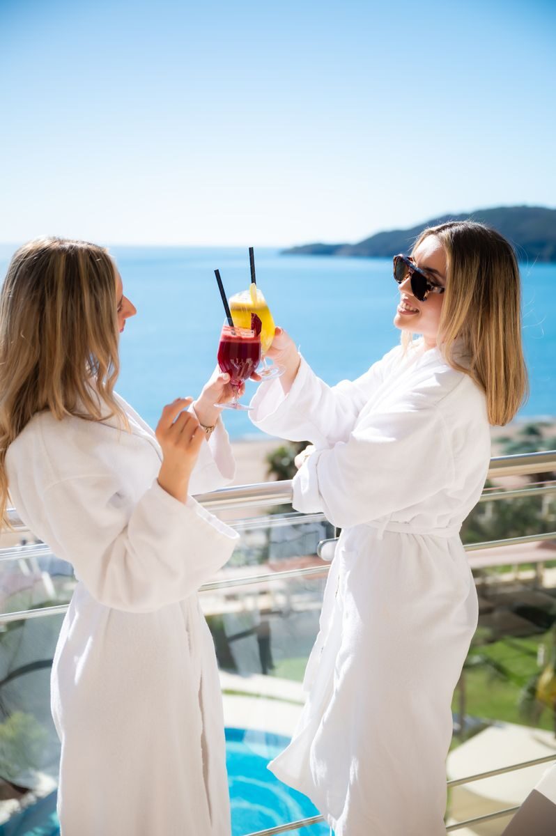 Two women in white robes toast with drinks on a sunny day by the sea, exuding relaxation and leisure.