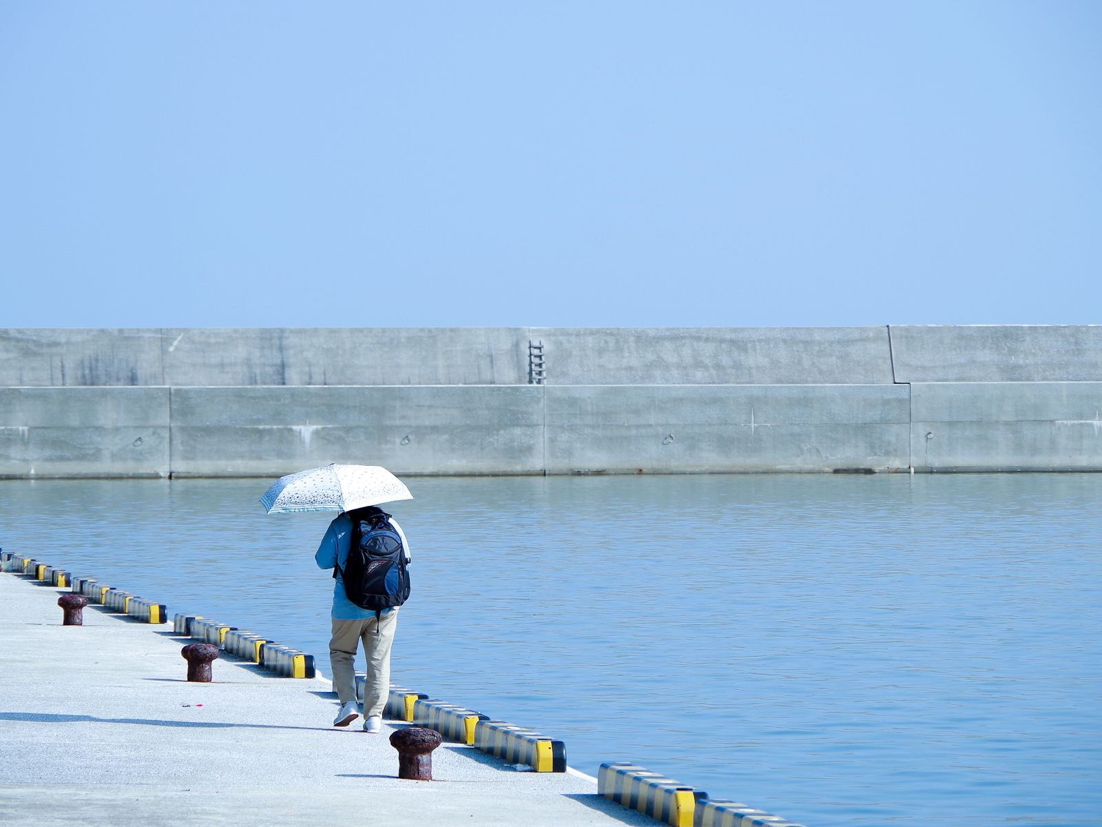 A solitary figure with umbrella walks along a sunny pier beside calm blue waters.
