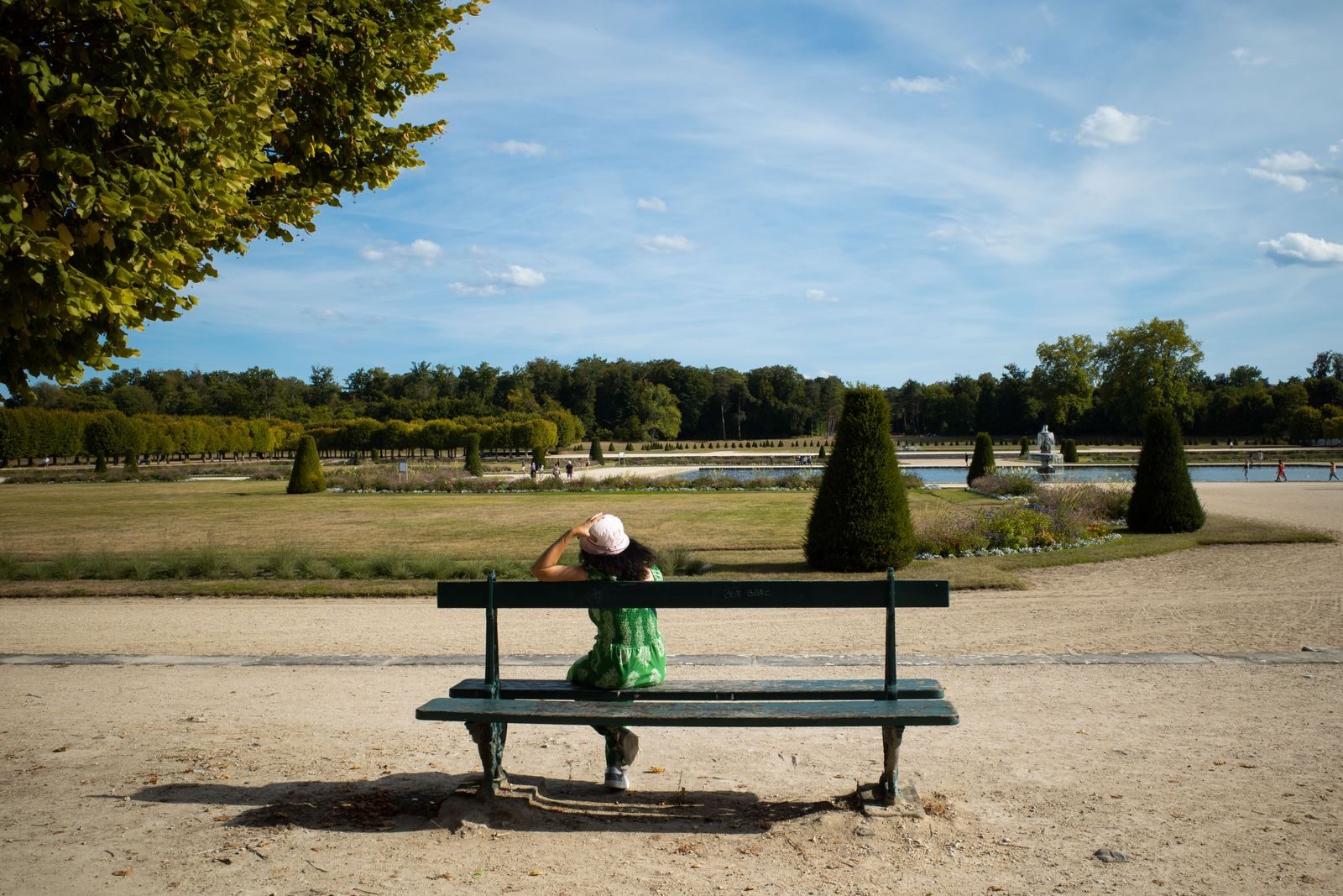 A woman in a green dress and hat relaxes on a bench in Fontainebleau Gardens, France, under a clear blue sky.