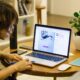 Woman working remotely from home with a laptop, pen, and plant on her desk, highlighting digital nomad lifestyle.