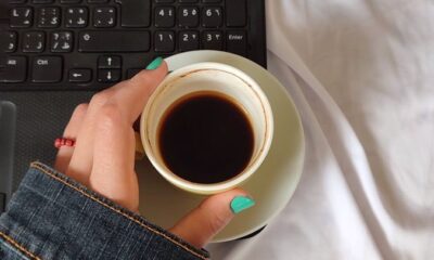 A person with turquoise nails holds a coffee cup near a laptop on a white bedspread.