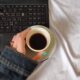 A person with turquoise nails holds a coffee cup near a laptop on a white bedspread.