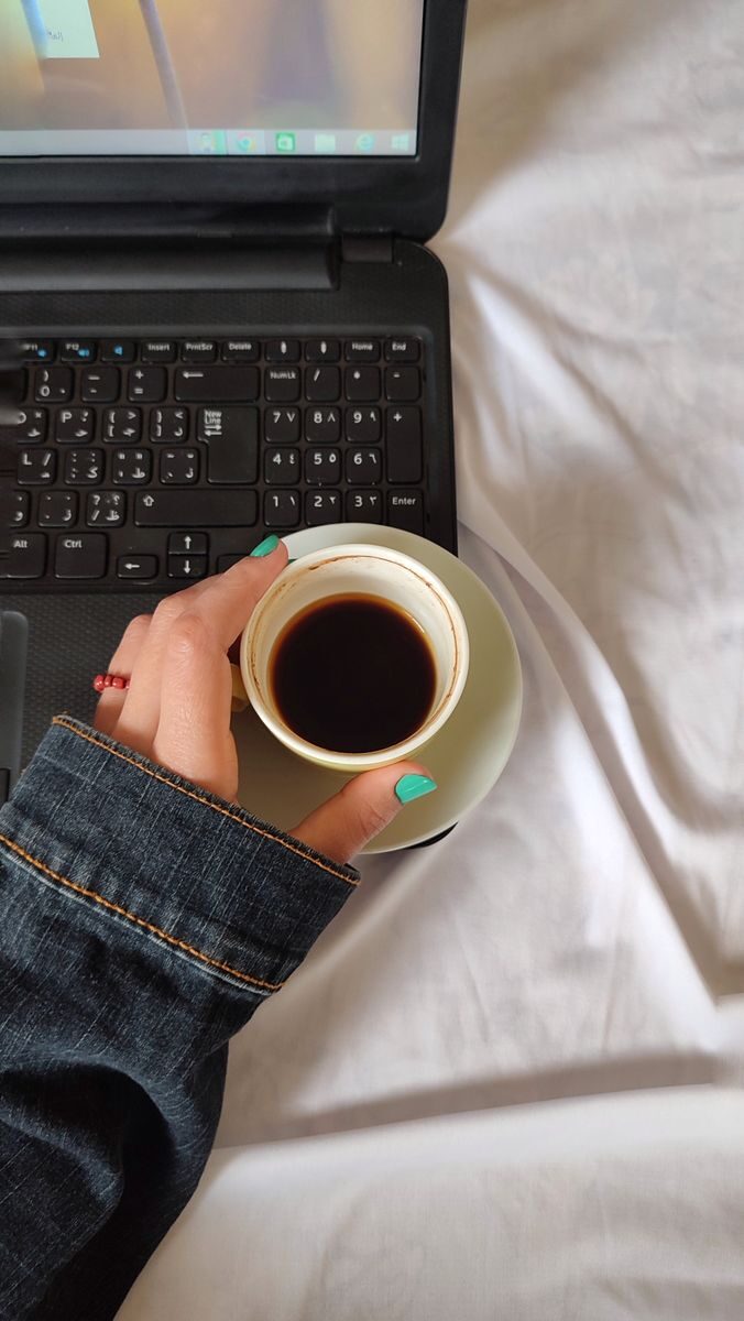 A person with turquoise nails holds a coffee cup near a laptop on a white bedspread.