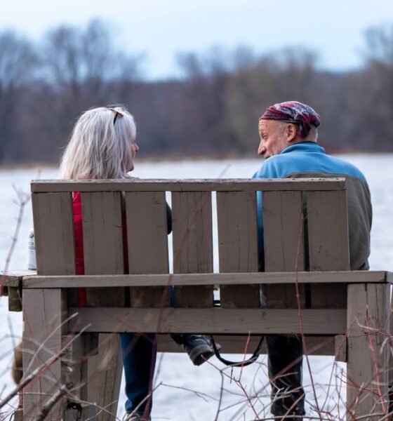 An elderly couple sits on a bench by a lake, enjoying nature in winter.