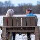 An elderly couple sits on a bench by a lake, enjoying nature in winter.