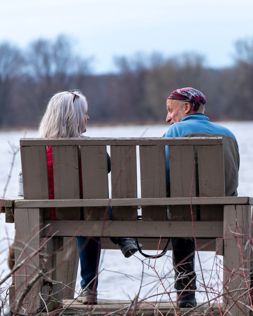 An elderly couple sits on a bench by a lake, enjoying nature in winter.