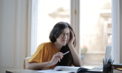 Ethnic female student in casual wear sitting at table with laptop and notebook and grimacing from migraine while doing homework during distance learning