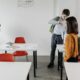 Students socializing in a classroom with red chairs and white desks, preparing to leave.