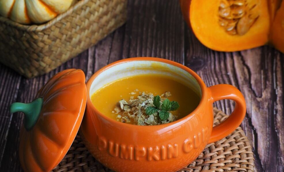 Close-up of fresh homemade pumpkin soup in an orange cup, surrounded by pumpkins.