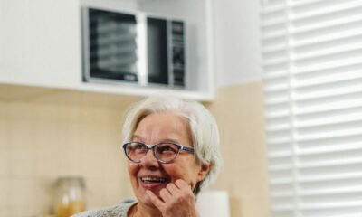 A cheerful elderly woman with glasses smiles warmly as she sits in a bright kitchen, adding a sense of comfort and homeliness.