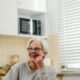 A cheerful elderly woman with glasses smiles warmly as she sits in a bright kitchen, adding a sense of comfort and homeliness.
