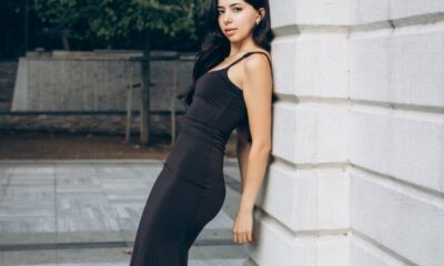 An elegant woman in a black dress poses by a stone building outdoors, showcasing fashion and style.