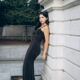 An elegant woman in a black dress poses by a stone building outdoors, showcasing fashion and style.