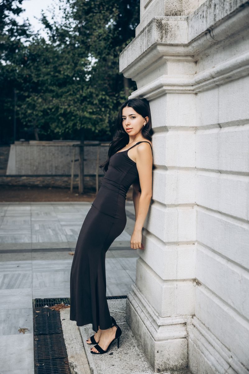 An elegant woman in a black dress poses by a stone building outdoors, showcasing fashion and style.