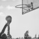 Two athletes compete for a basketball in an outdoor court in a dynamic black and white shot.