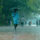 A man in blue walking under heavy rain with umbrella on a tree-lined path.
