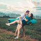 A couple romantically embracing in an open field with a vintage car and buildings in the background.
