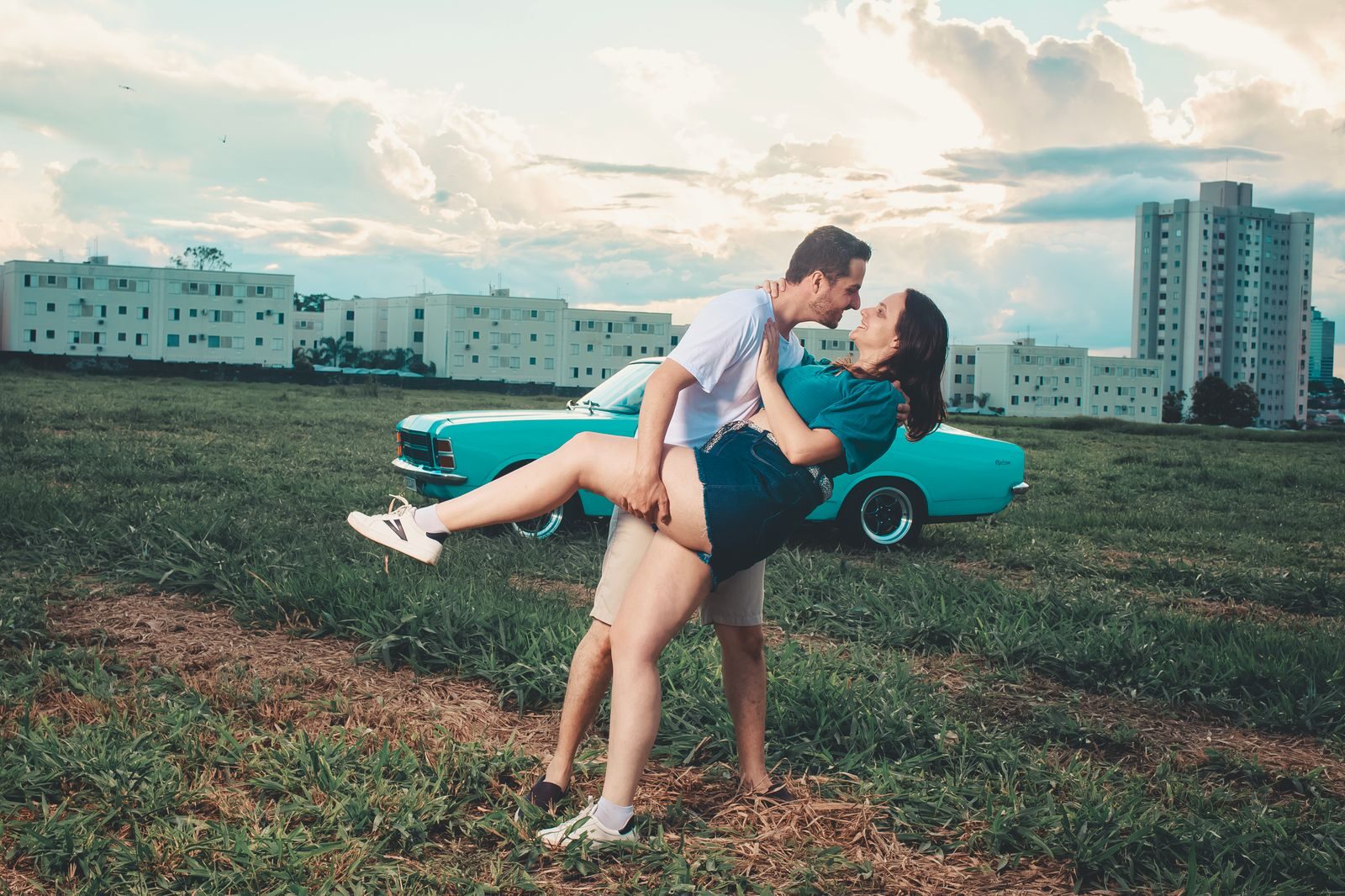 A couple romantically embracing in an open field with a vintage car and buildings in the background.