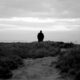 A solitary figure walking along a coastal path under a cloudy sky, captured in monochrome.