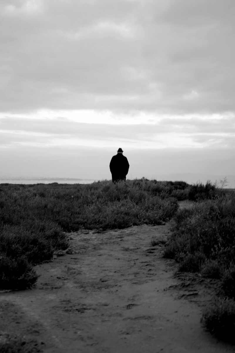 A solitary figure walking along a coastal path under a cloudy sky, captured in monochrome.