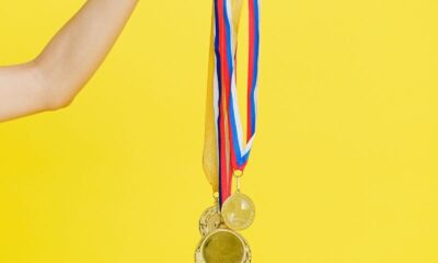 A hand holding multiple gold medals with colorful ribbons on a vibrant yellow background.
