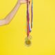 A hand holding multiple gold medals with colorful ribbons on a vibrant yellow background.