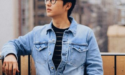 A stylish young man wearing glasses stands on a New York City balcony in a denim jacket.