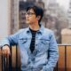 A stylish young man wearing glasses stands on a New York City balcony in a denim jacket.