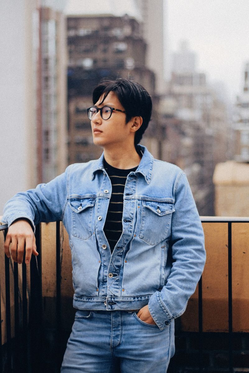 A stylish young man wearing glasses stands on a New York City balcony in a denim jacket.