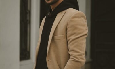 Man in beige coat posing outside an urban building, showcasing trendy fashion style.