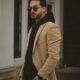 Man in beige coat posing outside an urban building, showcasing trendy fashion style.