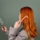 Red-haired woman using a comb while holding a mirror, focusing on hair care and beauty.