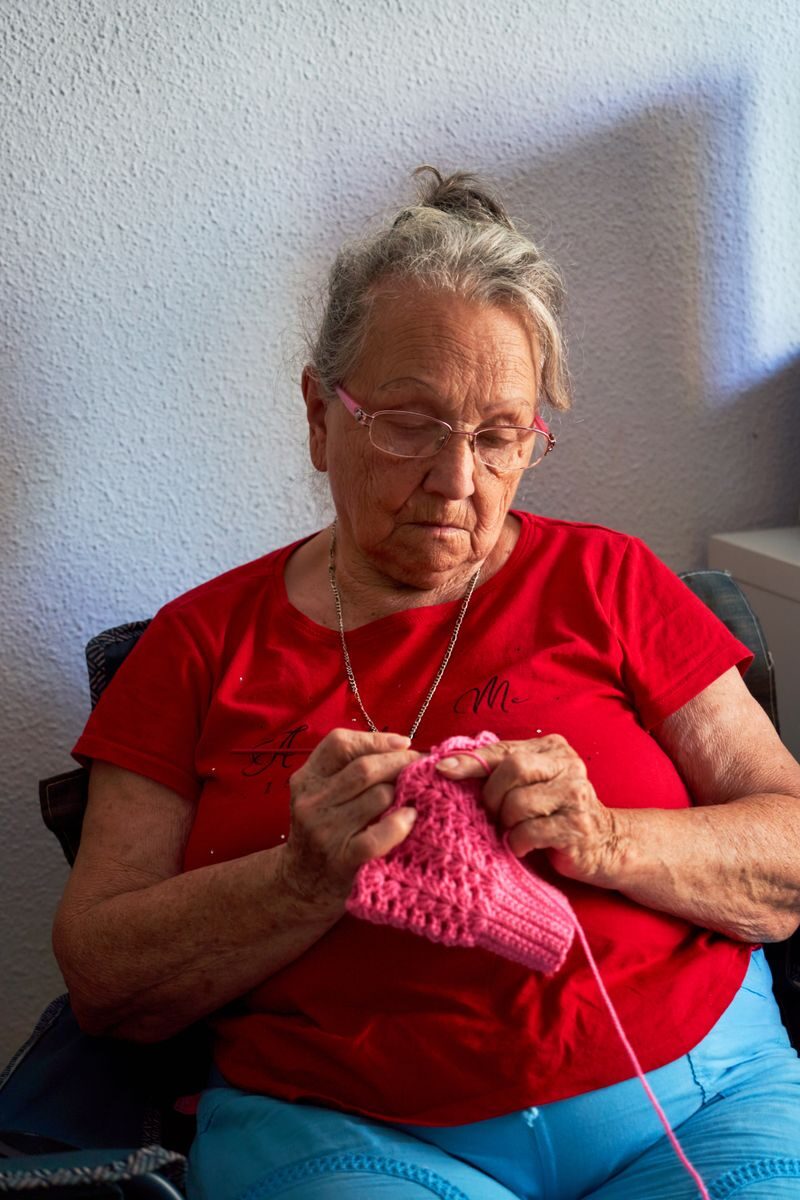 Senior woman in red shirt knitting pink yarn indoors, focusing on craft.