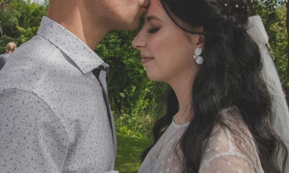 A groom kisses his bride's forehead during their outdoor wedding ceremony.